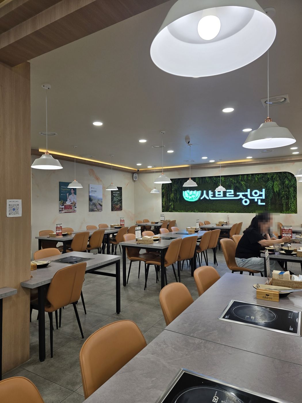 주황색 의자와 인덕션이 놓인 테이블이 줄지어 있는 실내, 벽면에 샤브르정원 네온 간판